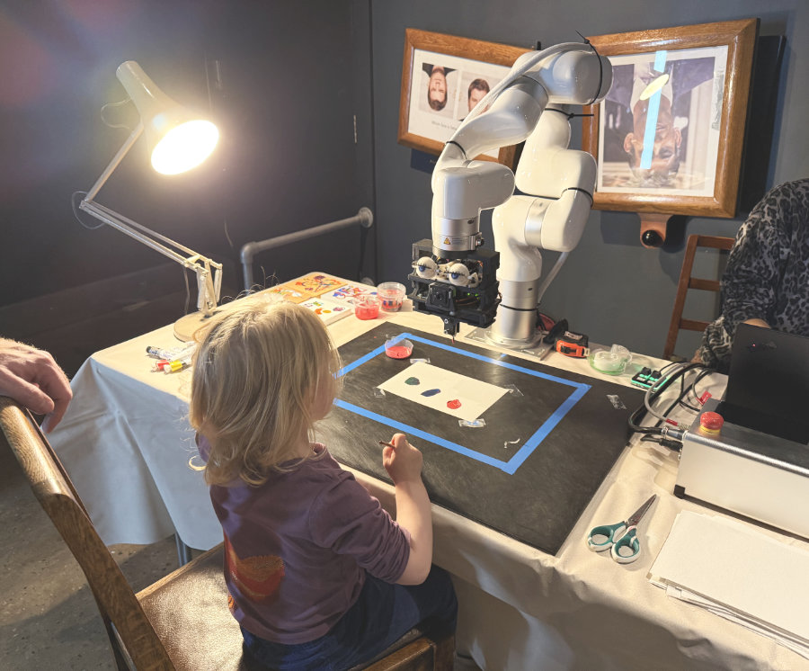 Ein kleines Kind sitzt an einem Tisch, während ein Roboterarm mit einer Kamera vor ihm eine Zeichnung anfertigt. Der Tisch ist mit Malutensilien und einer Schreibtischlampe ausgestattet, und im Hintergrund sind Bilder an der Wand zu sehen.