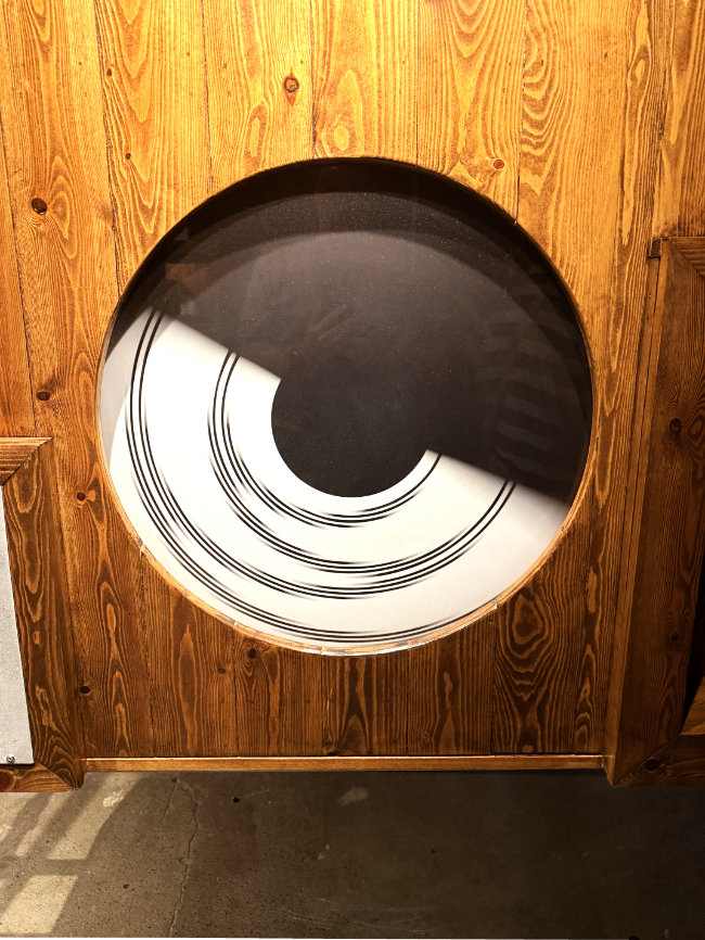 Eine runde, schwarz-weiße Kunstinstallation in einem hölzernen Rahmen, die einen spiralförmigen, abstrakten Entwurf zeigt.