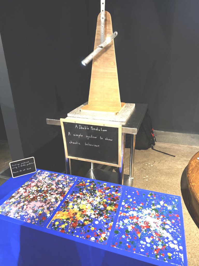 A double pendulum exhibit demonstrating chaotic behavior, with a wooden structure and colored artworks underneath.