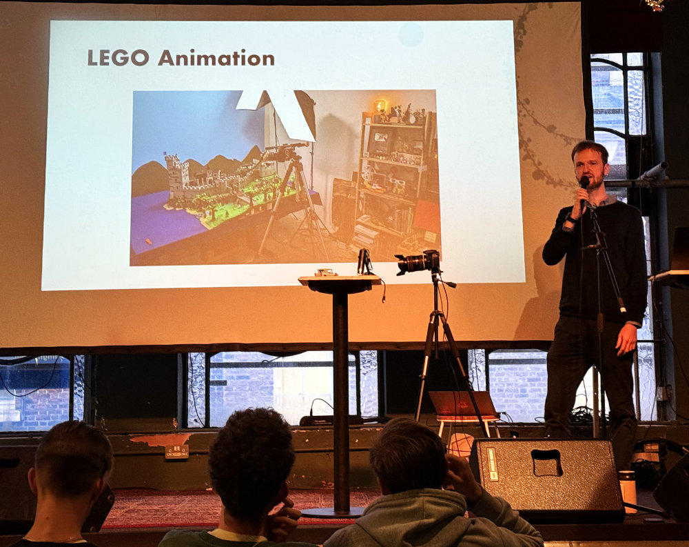 Ein Physiker präsentiert eine LEGO-Animation auf einer Bühne, während das Publikum ihn aufmerksam verfolgt. Im Hintergrund sind Schauspielrequisiten und eine Kamera zu sehen.