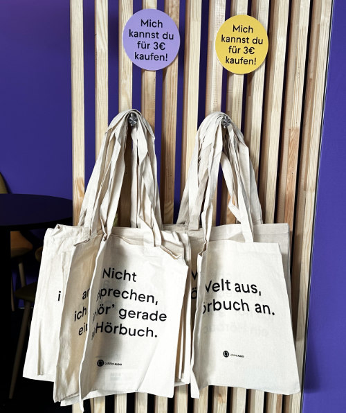 Überrascht von einer Auswahl an Stoffbeuteln, die auf einem Holzständer hängen, mit Aufschriften, die sich auf das Hören von Hörbüchern beziehen.