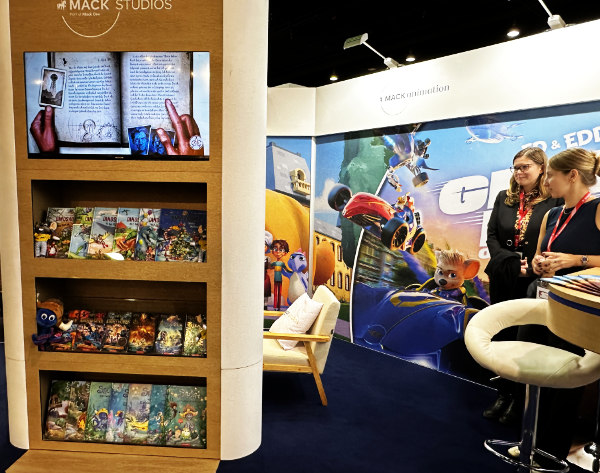 Ein Messestand von Mack Studios bei der Buchmesse mit einem Bildschirm, der ein offenes Buch zeigt, sowie einer Regale mit bunten Kinderbüchern und Figuren im Vordergrund.