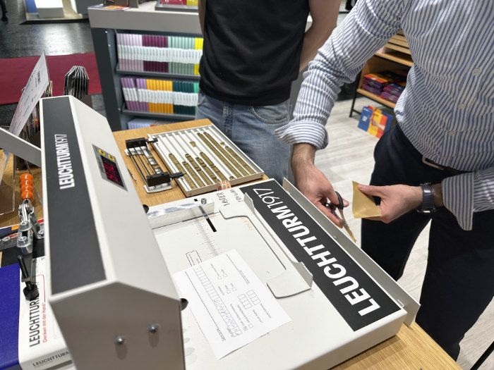 Ein Stand auf der Buchmesse mit zwei Personen, die an einem Tisch mit verschiedenen Schreibutensilien und Materialien arbeiten.