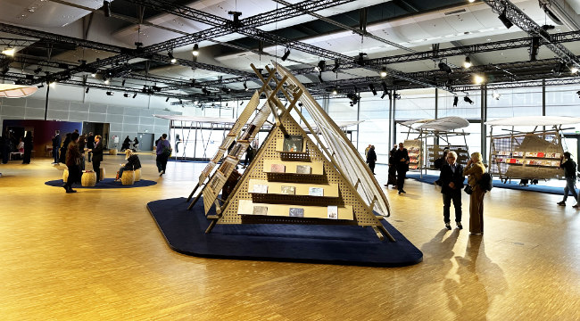 Ein modernes Ausstellungsdesign auf der Frankfurter Buchmesse mit mehreren Besuchern, die um eine pyramidenförmige Bücherpräsentation und Kunstinstallationen herumstehen.