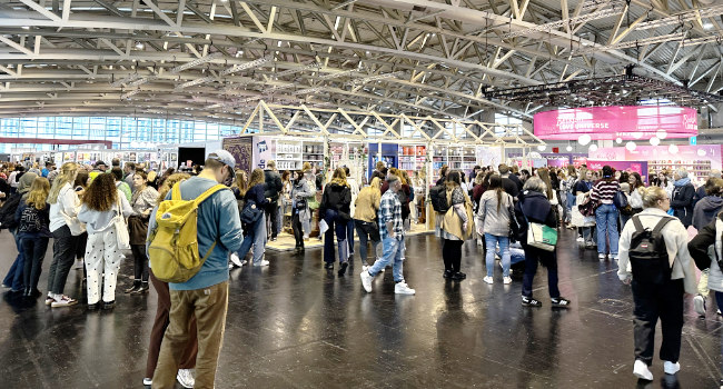 Blick auf die Frankfurter Buchmesse mit einer großen Menschenmenge, die zwischen Ausstellerständen wandert, in einer hellen Halle mit modernen architektonischen Elementen.