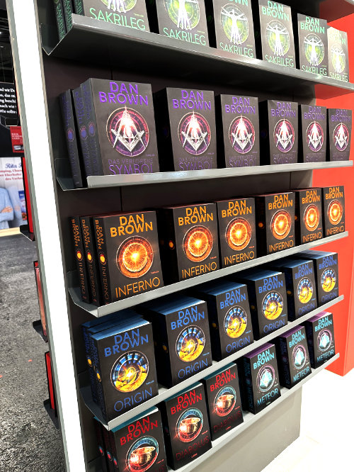 Regal mit Büchern von Dan Brown, darunter Titel wie 'Sakrileg', 'Inferno', 'Origin' und 'Meteor', gegen eine graue und rote Wand auf einem Buchmesse-Stand.