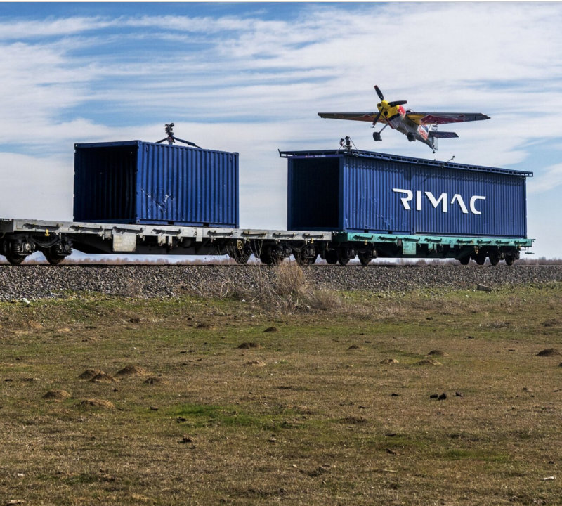Ein Flugzeug fliegt über zwei blaue Frachtenzüge mit dem Schriftzug 'RIMAC' auf den Containern, während sie auf Schienen stehen.